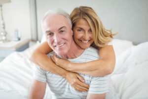 Older couple smiling on a neatly made bed, with the woman’s arms wrapped around the man.